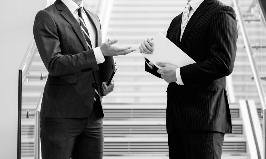 スーツ姿のビジネスパーソンが資料を持って立ち話をする様子
